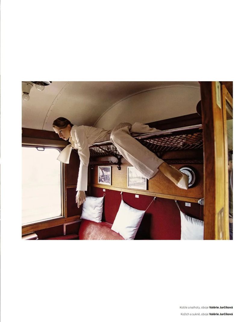 Indoor shot of a person in a white suit lying on a sleeping rack above a red seat inside a train car with a window to the outside.