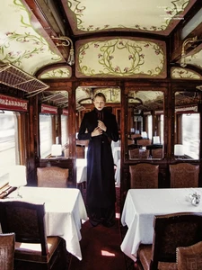 Model standing inside a vintage wood-paneled train dining car wearing a dark long coat for Harper's Bazaar Czech Republic