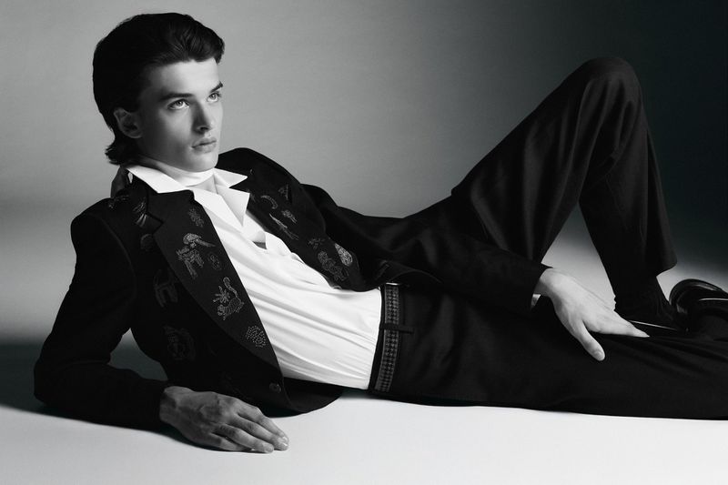 Portrait of a young adult male model in a black suit and white shirt lying on his side against a gradient gray backdrop in a photo studio.