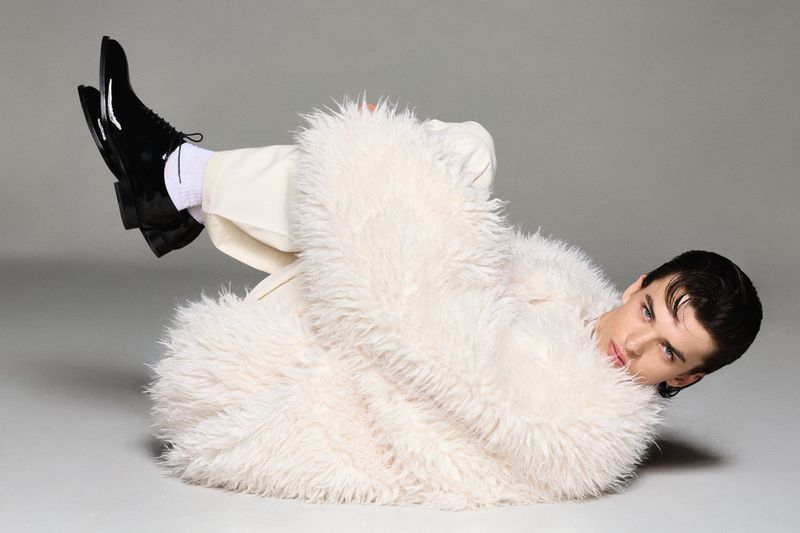 Adult male fashion model poses on gray backdrop wearing white pants, black patent leather dress shoes and a fuzzy white coat.