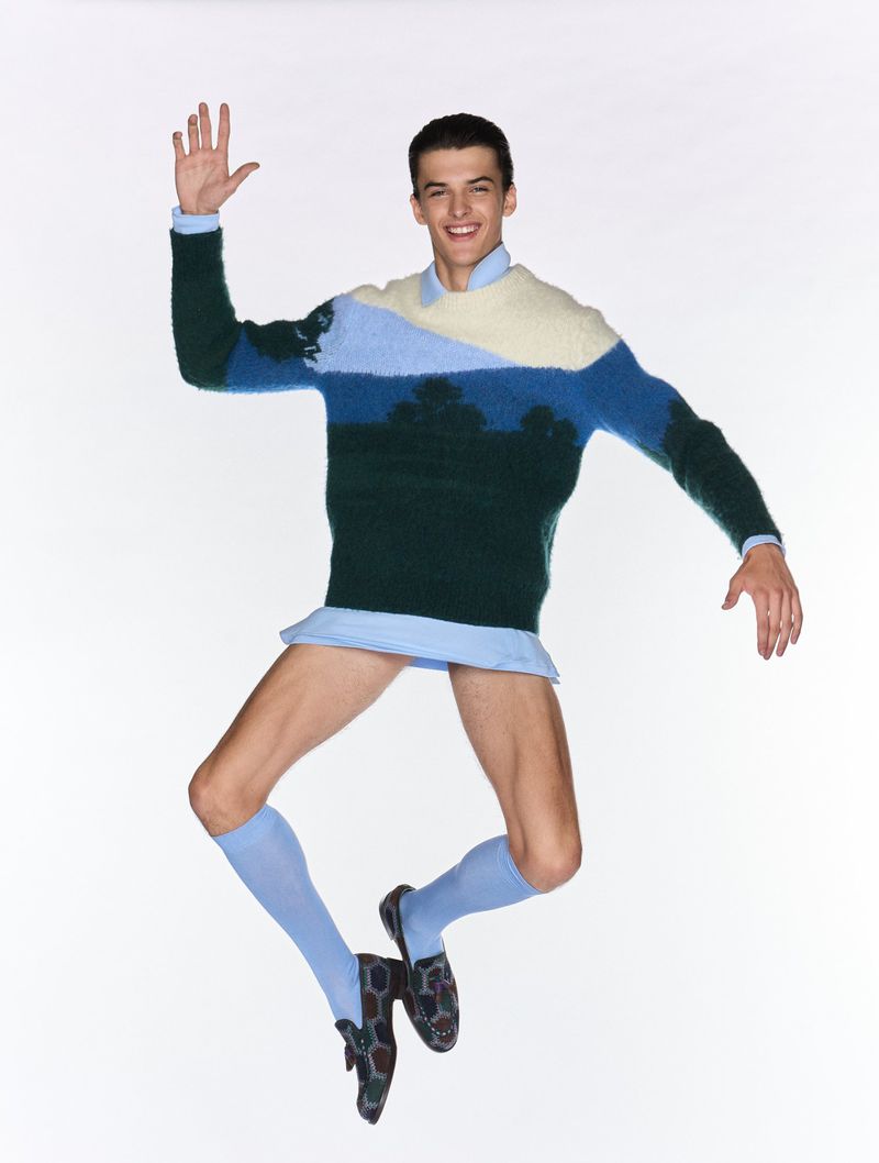 Fashion shot of a smiling man in a patterned sweater, light blue socks, and loafers, jumping in a white photo studio, raising his hand.