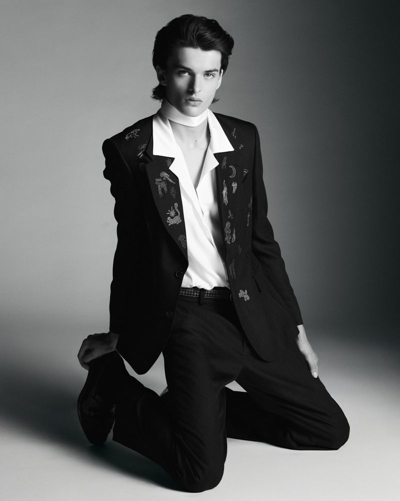 Portrait of a young adult male in a suit kneeling, featuring black and white tones, studio lighting, and a fashion-forward aesthetic.