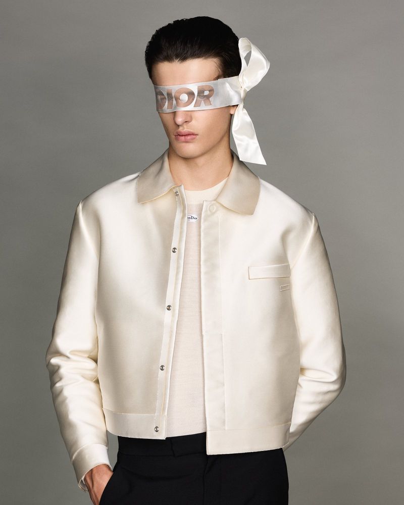 Portrait of a male fashion model wearing a white cropped jacket and a Dior-branded blindfold, posing against a neutral grey background.