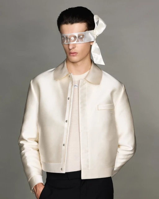 Portrait of a male fashion model wearing a white cropped jacket and a Dior-branded blindfold, posing against a neutral grey background.