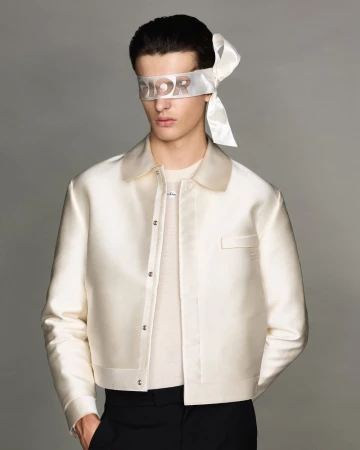Portrait of a male fashion model wearing a white cropped jacket and a Dior-branded blindfold, posing against a neutral grey background.