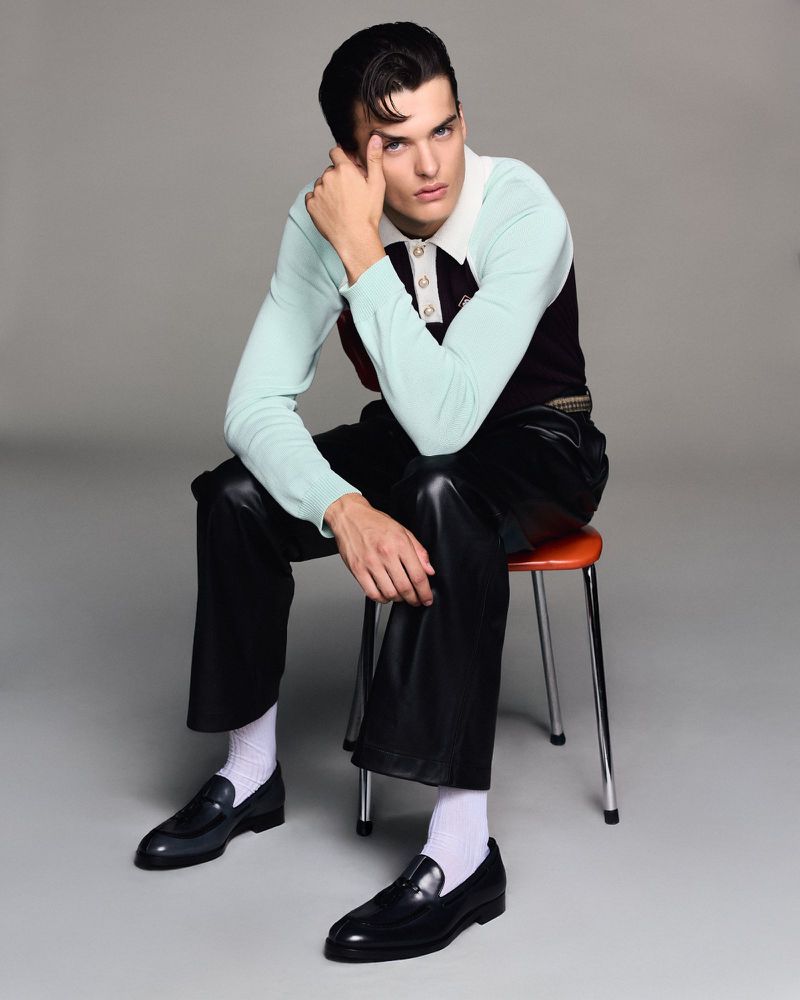 Adult male sits on a stool in a photo studio. He is wearing leather pants, dress shoes, and a collared top with white socks.