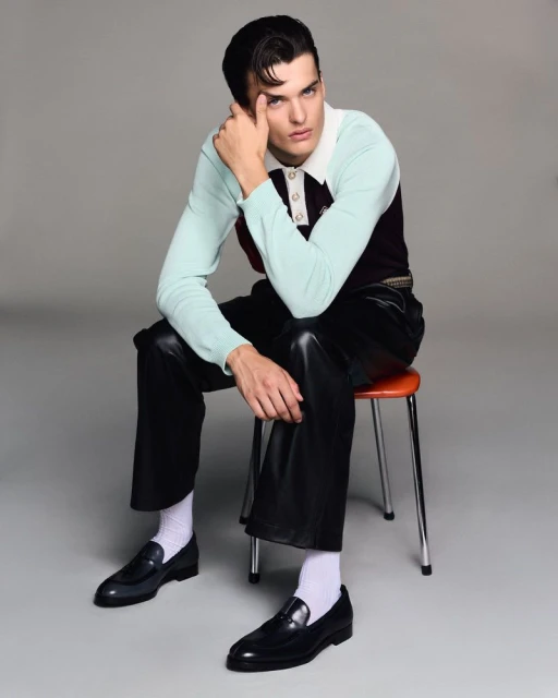 Adult male sits on a stool in a photo studio. He is wearing leather pants, dress shoes, and a collared top with white socks.