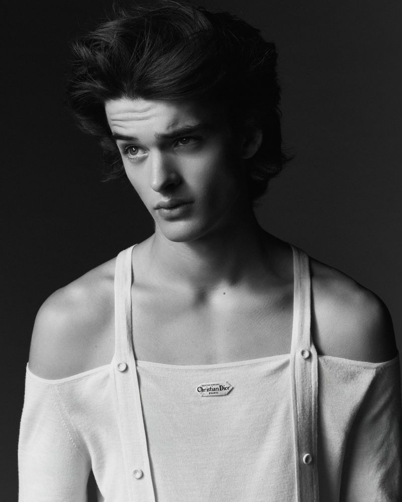 Monochrome portrait of a young adult male with dark hair, wearing an off-the-shoulder white top, posing against a dark background.