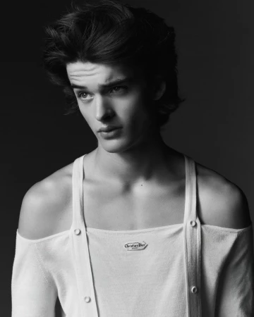 Monochrome portrait of a young adult male with dark hair, wearing an off-the-shoulder white top, posing against a dark background.