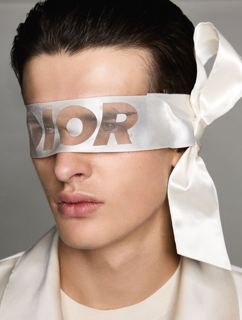 Close-up of a young adult male model wearing a DIOR-branded white silk blindfold over his eyes in a beauty and fashion shot.