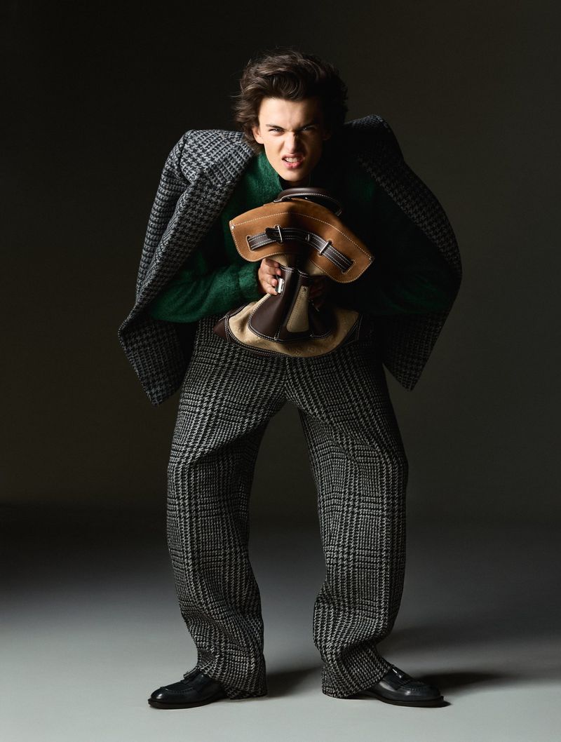 A young adult man wearing tweed pants and a coat, carrying a brown bag, poses with an intense expression in a studio setting.