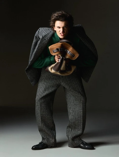 A young adult man wearing tweed pants and a coat, carrying a brown bag, poses with an intense expression in a studio setting.