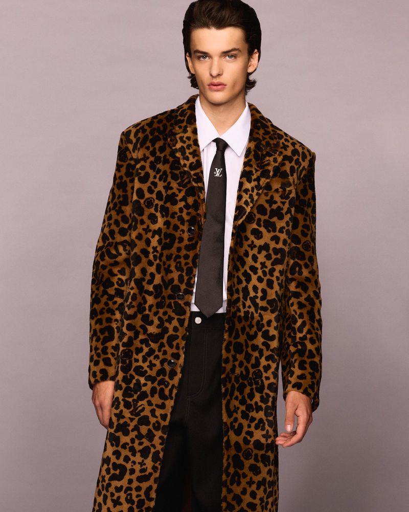 Man in a cheetah-print coat, white dress shirt, and dark tie poses in front of a studio backdrop looking into the camera.