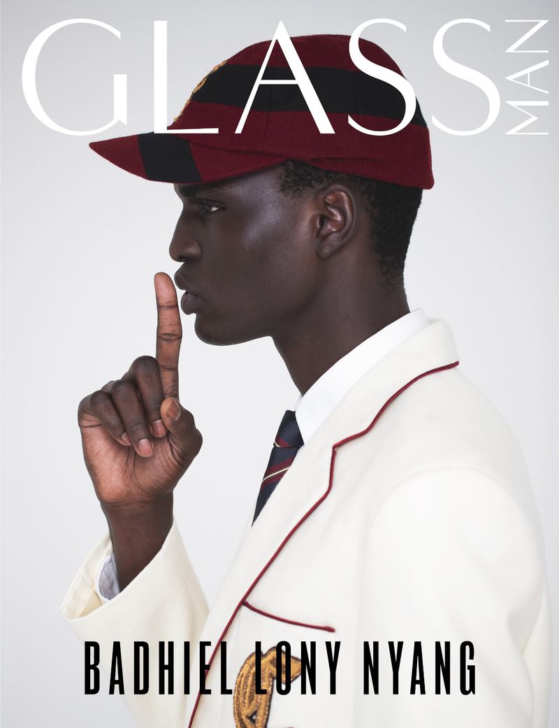 Fashion magazine cover featuring a Black male model in a jacket and cap holding a finger to his lips, posing against a neutral backdrop.