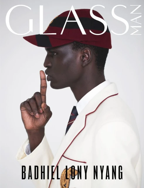 Fashion magazine cover featuring a Black male model in a jacket and cap holding a finger to his lips, posing against a neutral backdrop.