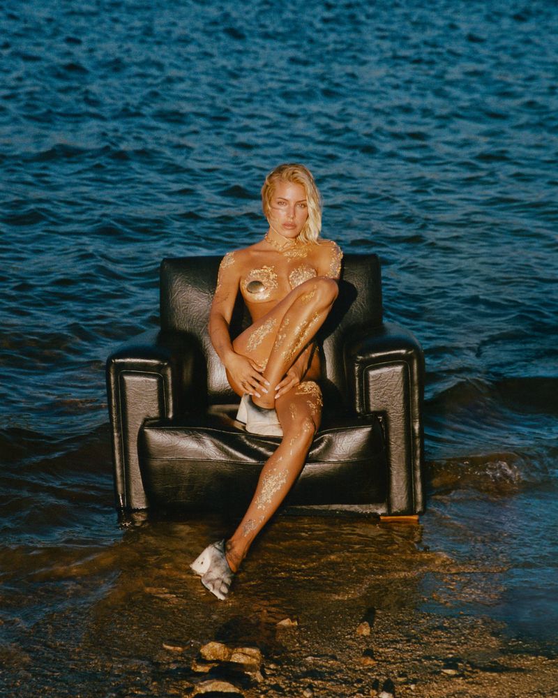 Woman with golden body art sits in armchair on shoreline of wavy blue ocean, wearing mismatched silver shoes in outdoor setting.