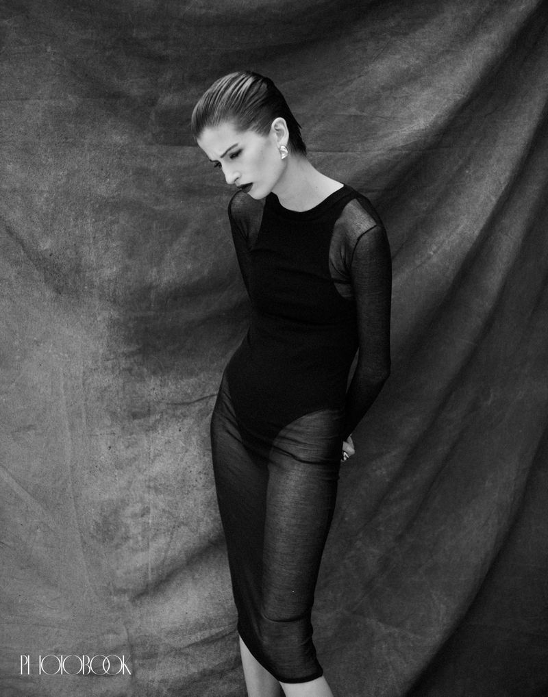 Woman in a black outfit posing against a textured grey backdrop. Black and white fashion shot with studio lighting and elegant style.