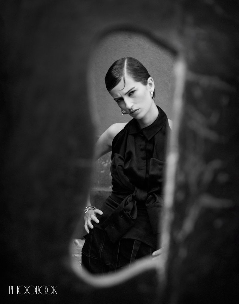 Black-and-white portrait of a young woman with slicked-back hair, wearing a dark satin top, hands on lap, shot through abstract hole.