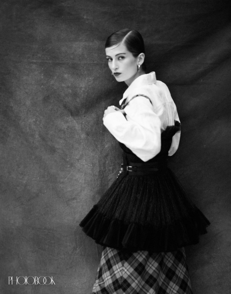 Black and white portrait of a woman in a white collared shirt, a short black skirt, and a plaid skirt, set against a neutral backdrop.