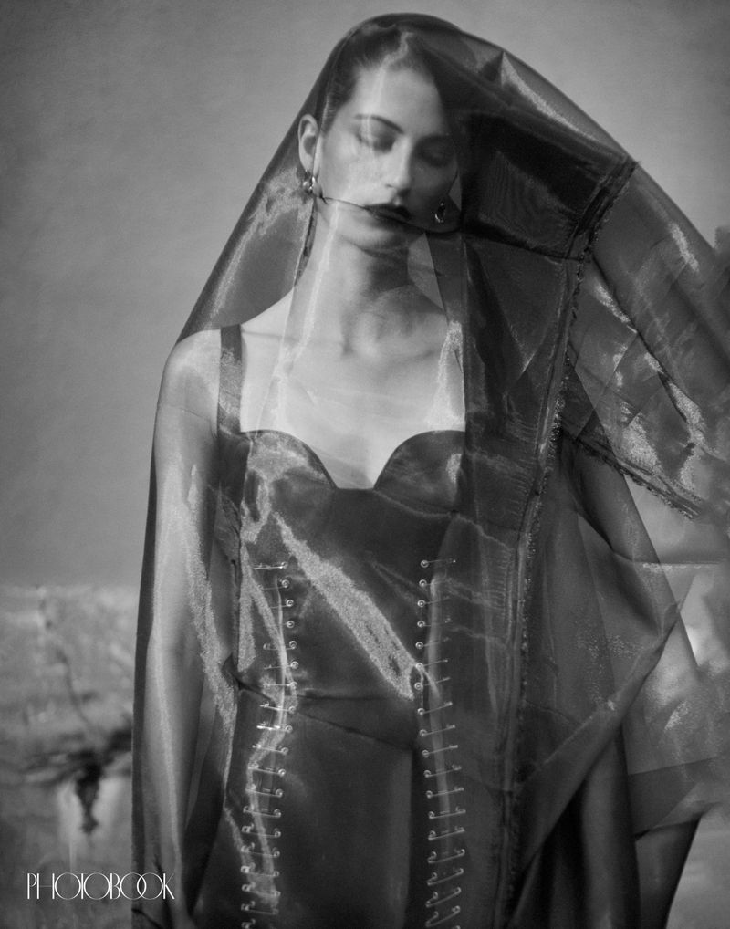 Monochrome portrait of a woman with eyes closed, wearing a structured bodice and sheer veil against a neutral textured backdrop.