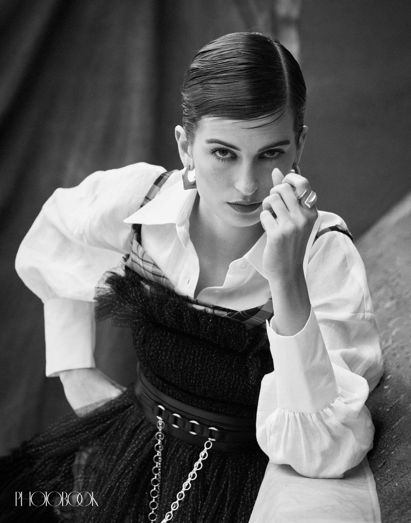 Black and white portrait of a brunette adult woman in a collared shirt and dress, accented by jewelry, with draped fabric behind.