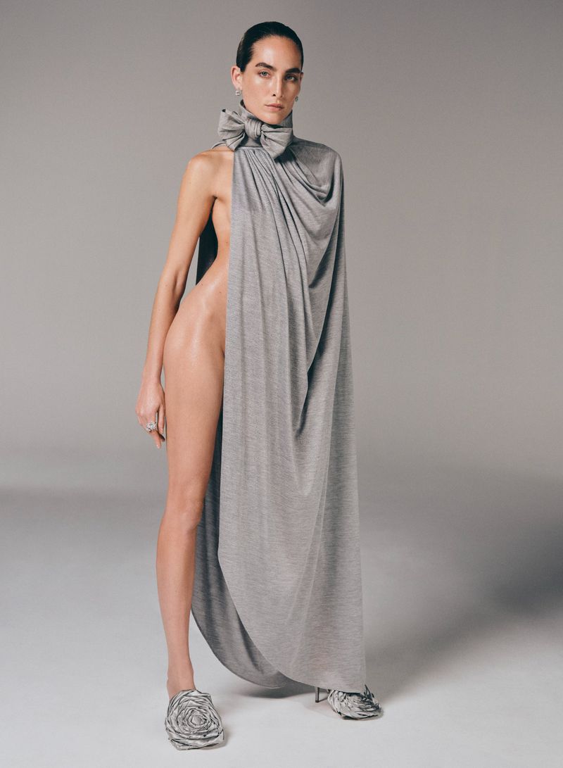 A model poses in a draped grey gown and sculptural shoes against a neutral background, showcasing a blend of fashion and art.