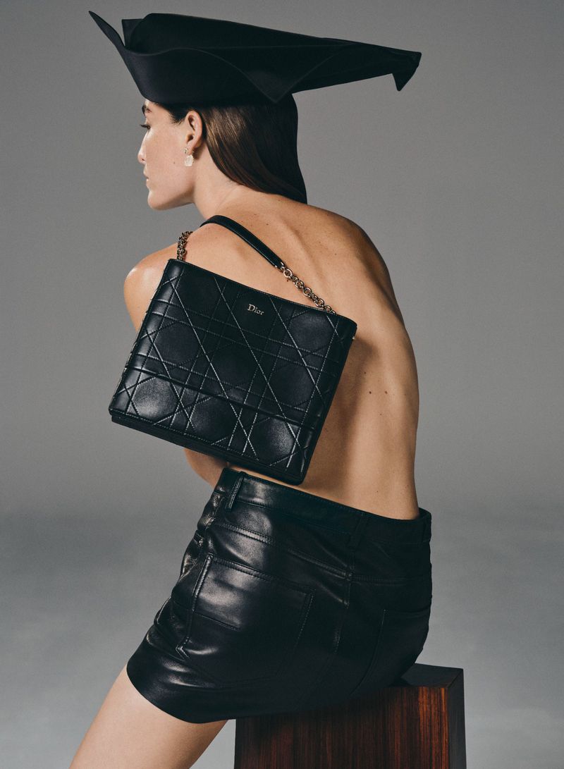 A fashion shot features a woman with a black shoulder bag and leather skirt. She wears a black headpiece and sits against a grey backdrop.