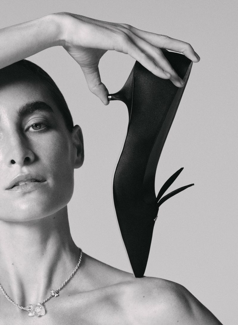 Black stiletto heel held by a fair-skinned woman with short hair and necklace, against a neutral studio background, fashion beauty shot.