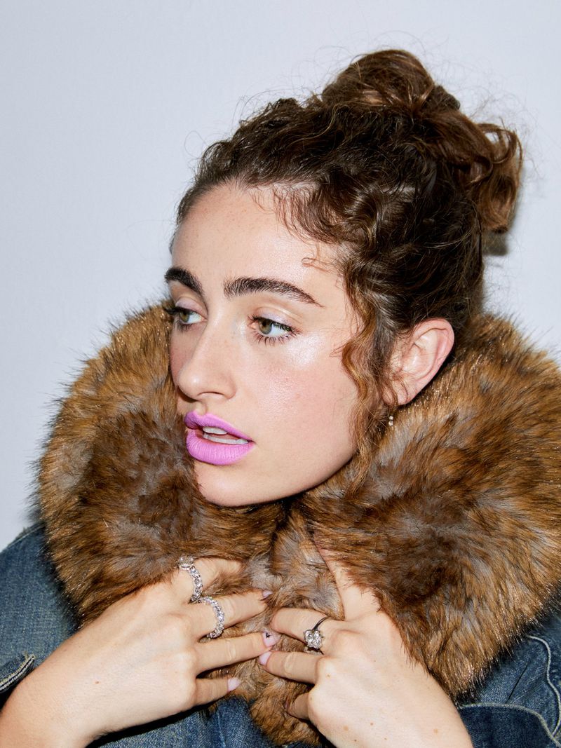 Portrait of a curly-haired woman with pink lipstick wearing a fur-collared denim jacket and statement rings against a plain white background.