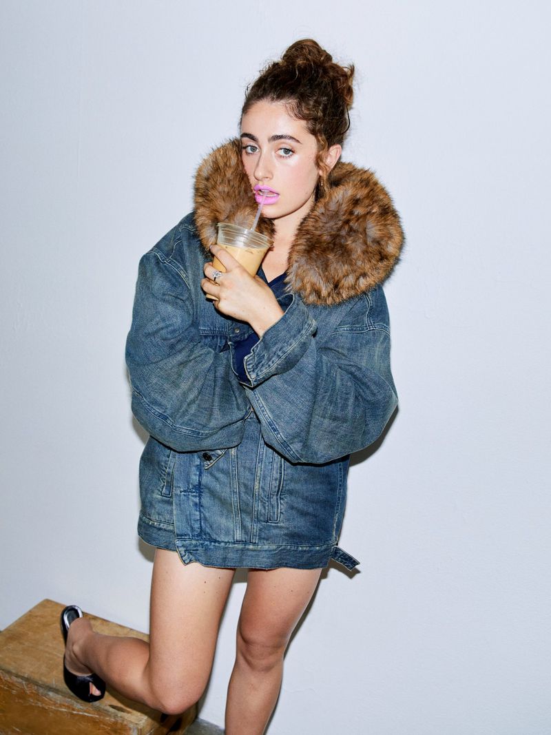 Young woman poses in denim jacket with fur collar, drinking from a clear plastic cup. White backdrop, casual fashion, and style.