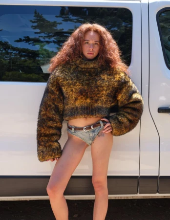 Model with long curly red hair wearing a fuzzy sweater and denim underwear leaning against a white vehicle