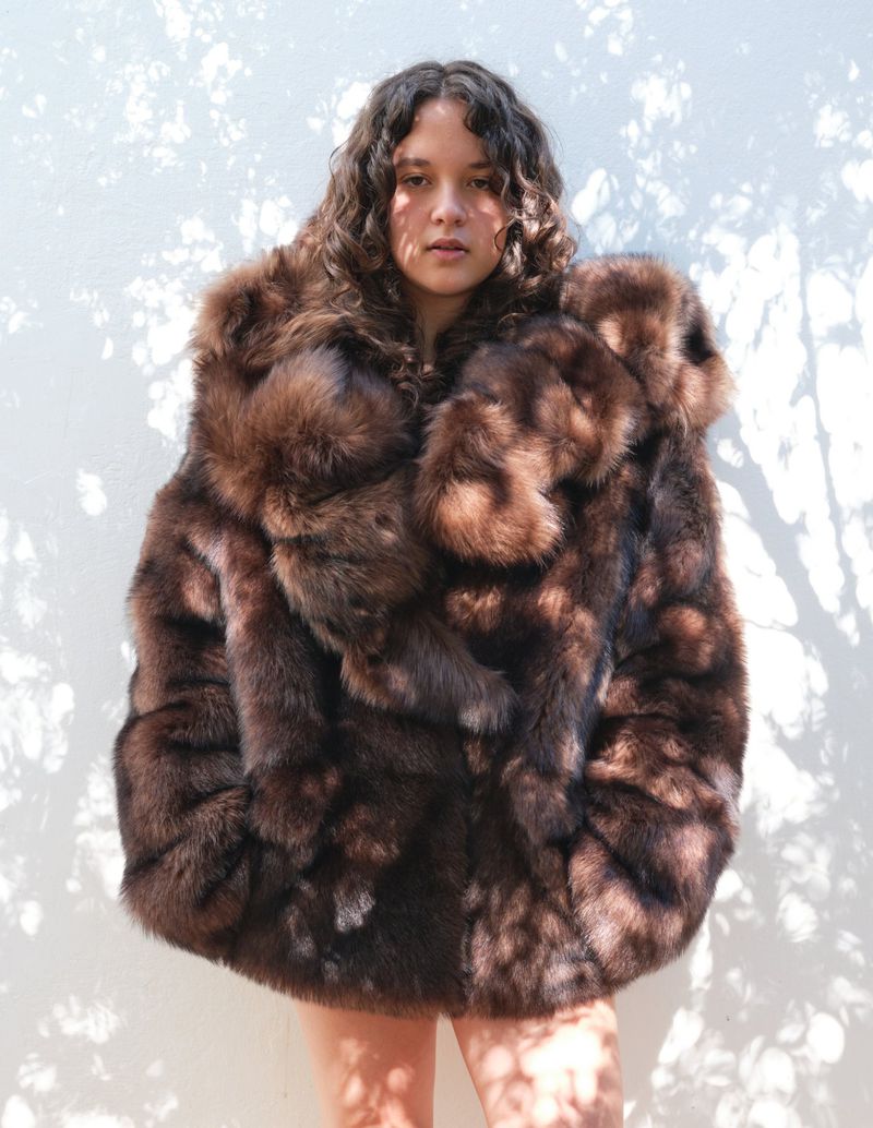 A young woman with curly hair poses in front of a white wall, wearing a large brown fur coat in an outdoor setting.