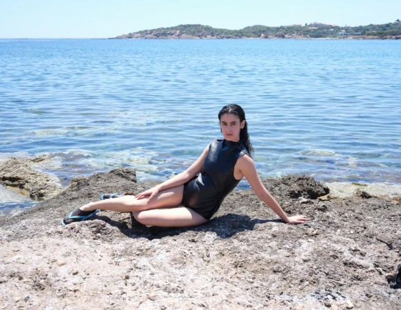Woman wearing a dark swimsuit reclining on rocky shore by the ocean for Double Magazine editorial photographed by Liv Liberg