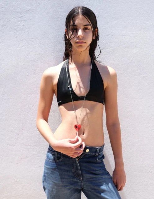 Young woman in a black halter top and jeans poses against a white wall with a heart necklace, creating a fashion portrait.