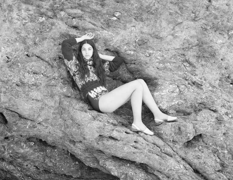 Black and white portrait of a young adult woman with long hair posing on a rocky surface in a sweater and shoes.