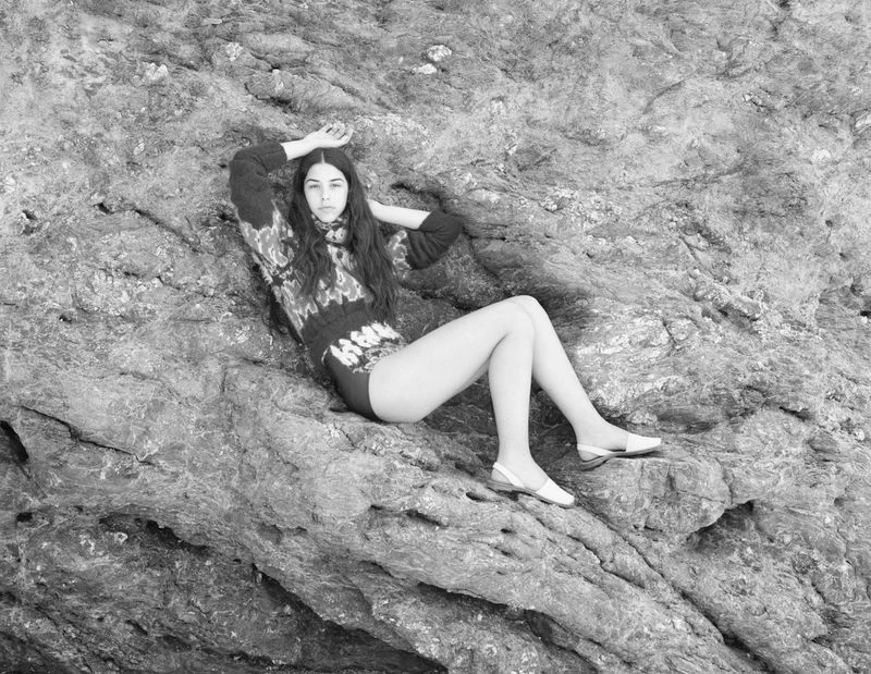 Black and white portrait of a young adult woman with long hair posing on a rocky surface in a sweater and shoes.