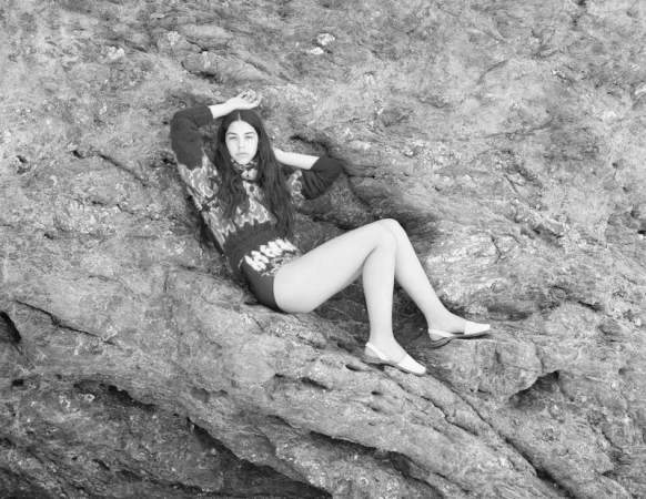 Black and white portrait of a young adult woman with long hair posing on a rocky surface in a sweater and shoes.