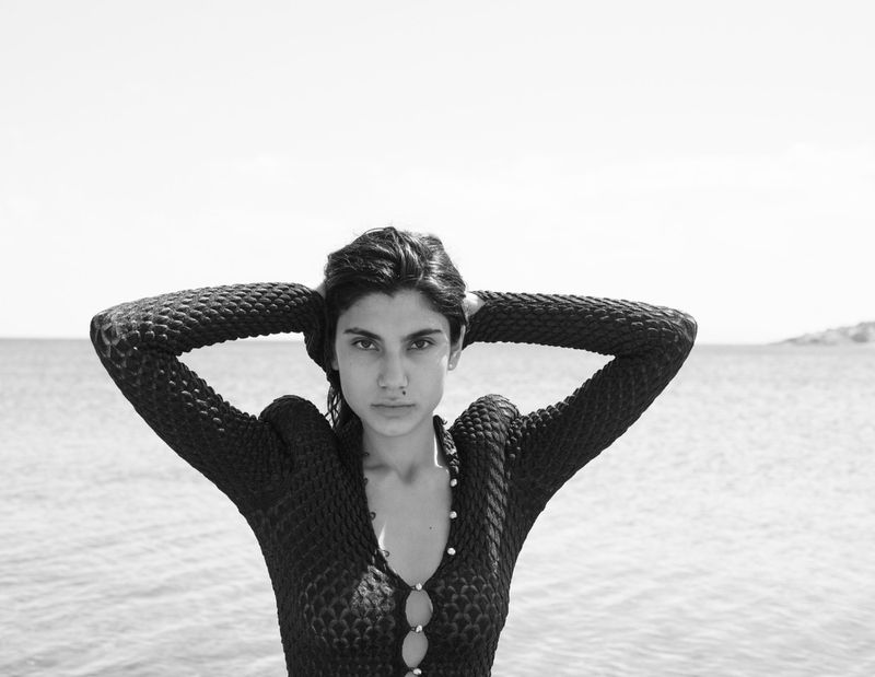 Monochromatic portrait of a woman with her hands behind her head, posing against a bright sky and the sea.