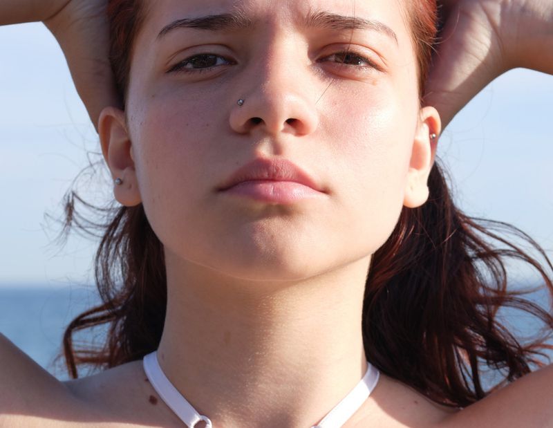 Close-up portrait of a red-haired woman with hands behind her head, set against a blue sky and ocean backdrop on a sunny day.