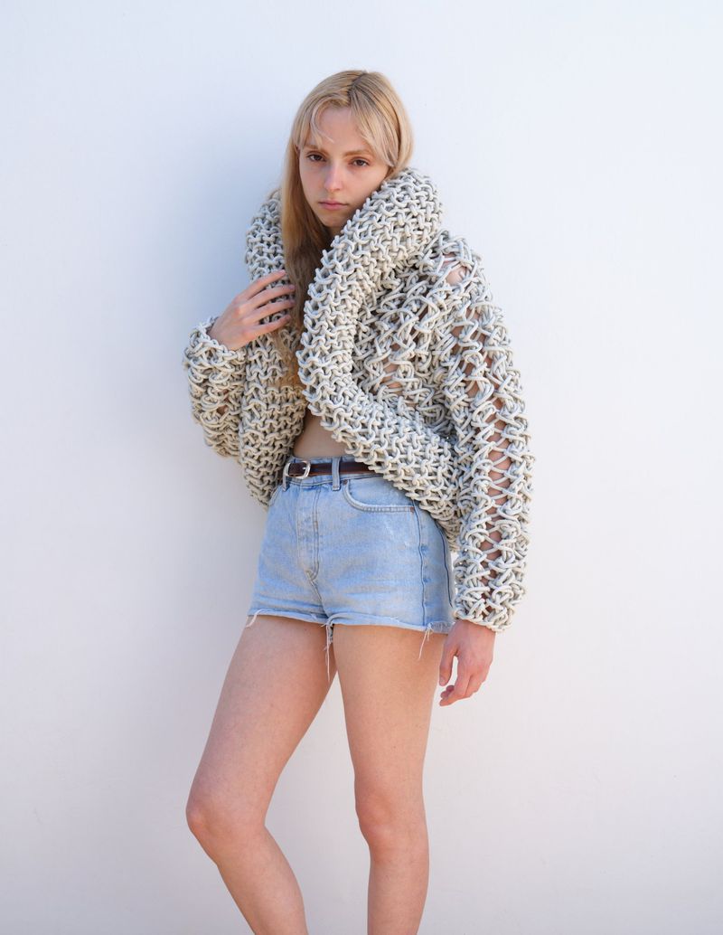 A fair-skinned young woman poses against a white wall, wearing a crocheted sweater and denim shorts in a brightly lit indoor setting.