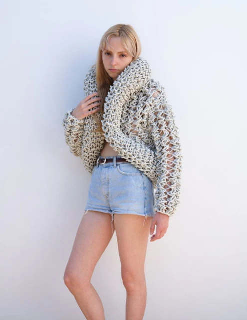 A fair-skinned young woman poses against a white wall, wearing a crocheted sweater and denim shorts in a brightly lit indoor setting.