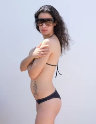 Woman standing in profile wearing oversized shield sunglasses and a black bikini against a pale seamless background.