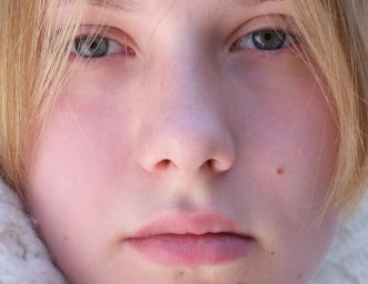 Extreme close-up portrait of a young blonde person with blue-grey eyes, freckles, and a neutral expression.