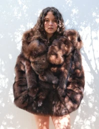 Model wearing an oversized brown fur coat standing against a sun-dappled white wall.