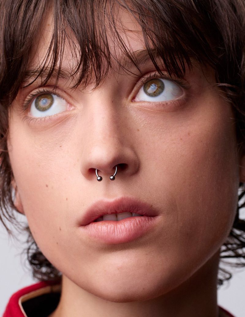 Close-up portrait of a young adult woman with a septum piercing, light skin, brown hair, and green eyes, looking upward in a neutral indoor setting.