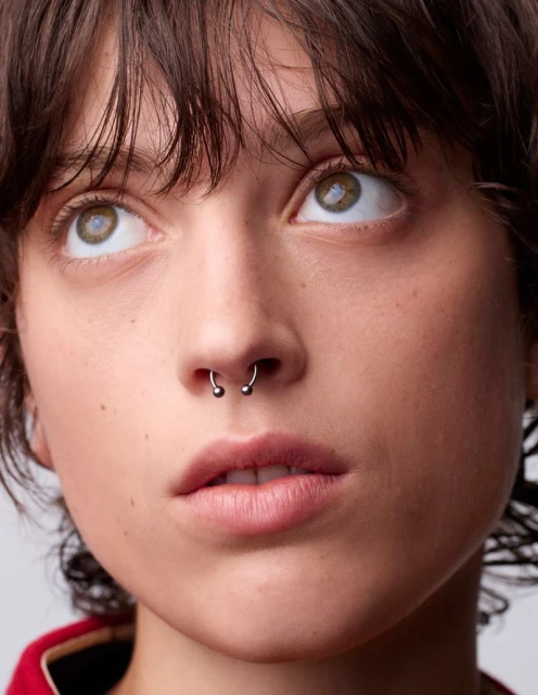 Close-up portrait of a young adult woman with a septum piercing, light skin, brown hair, and green eyes, looking upward in a neutral indoor setting.