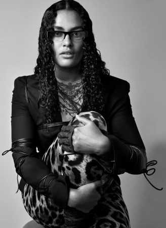 Model with long dark curly hair wearing glasses and a black jacket over leopard print pants, holding a patterned object against a grey background.
