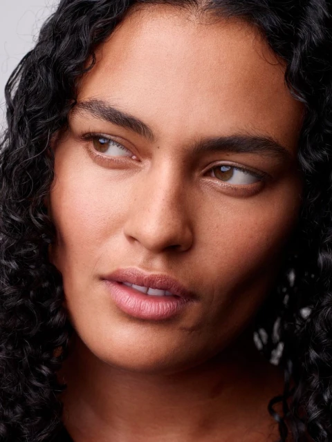 Close-up portrait of a woman with curly dark hair looking off to the side against a neutral backdrop with natural light. Beauty shot.