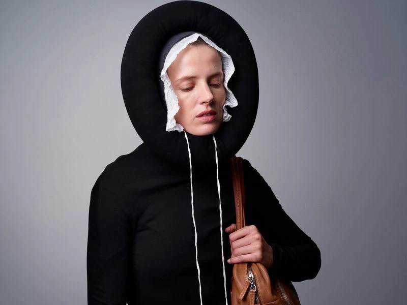 Woman in avant-garde black outfit with a padded hood and white ruff, holding a brown purse against a neutral gray backdrop.