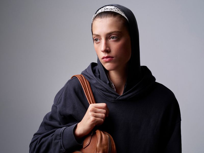 Portrait of an adult female wearing a dark hoodie with a white headband, looking away from the camera, and carrying a brown leather bag.
